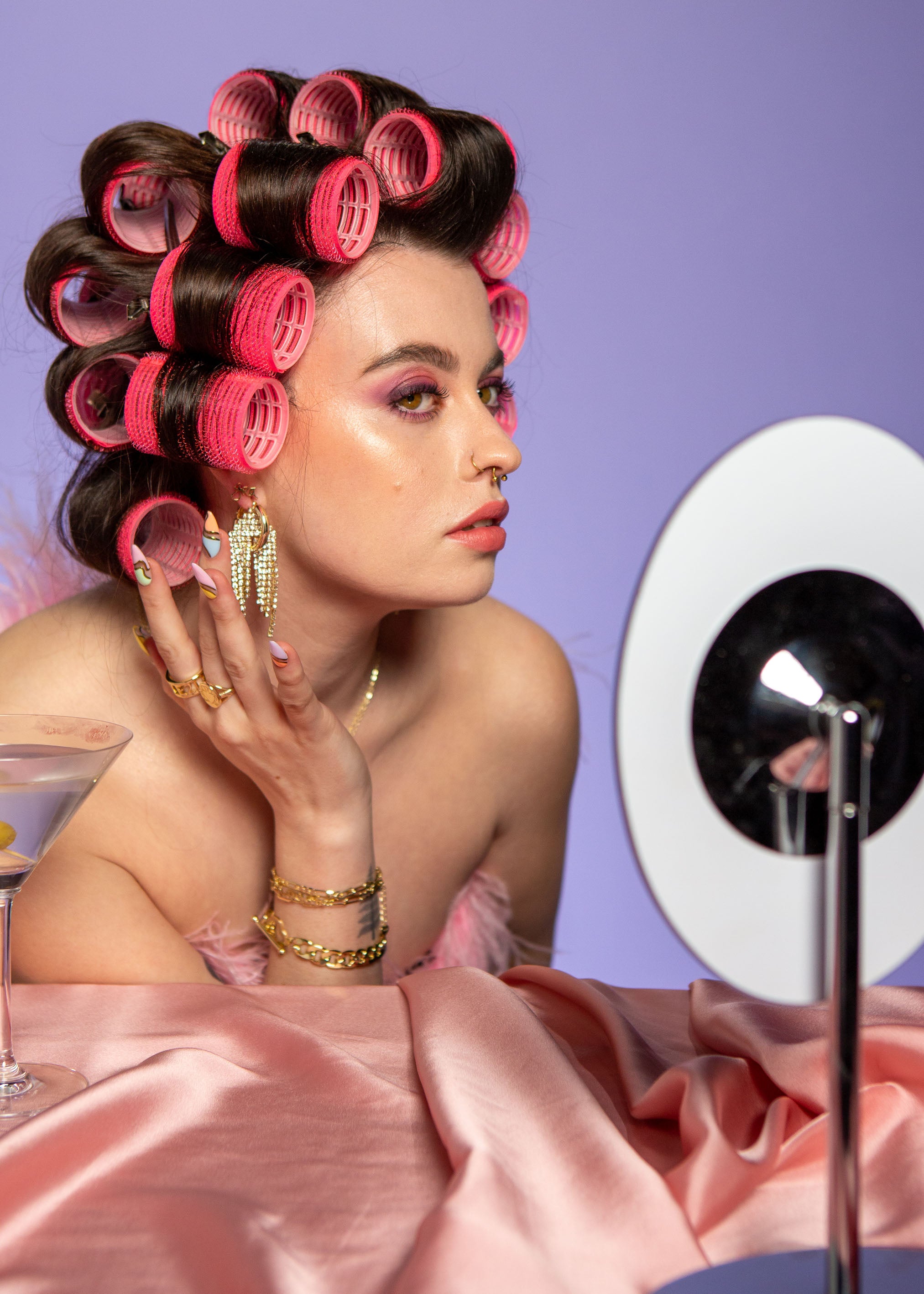 Woman with curled hair rollers in bright pink, wearing earrings, seated beside a vanity mirror