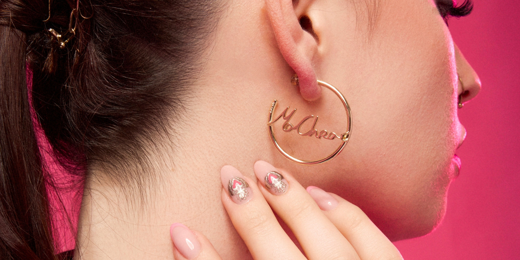 Woman wearing a gold hoop earring with script writing, close up with manicured nails and pink lighting